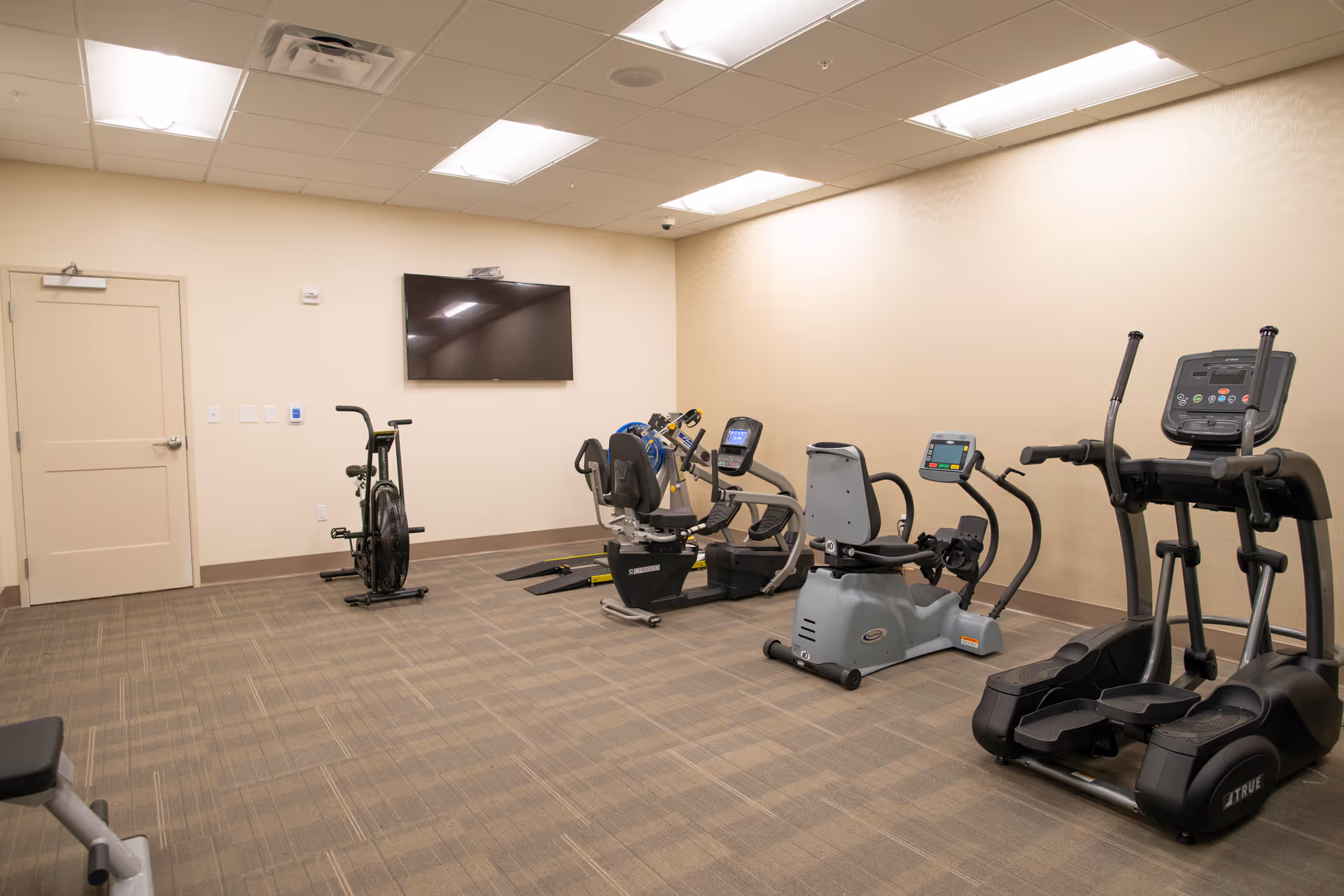 A small fitness room with exercise equipment including stationary bikes, a recumbent bike, and an elliptical machine. A flat-screen TV is mounted on the wall, and the room has beige walls and carpeted flooring.