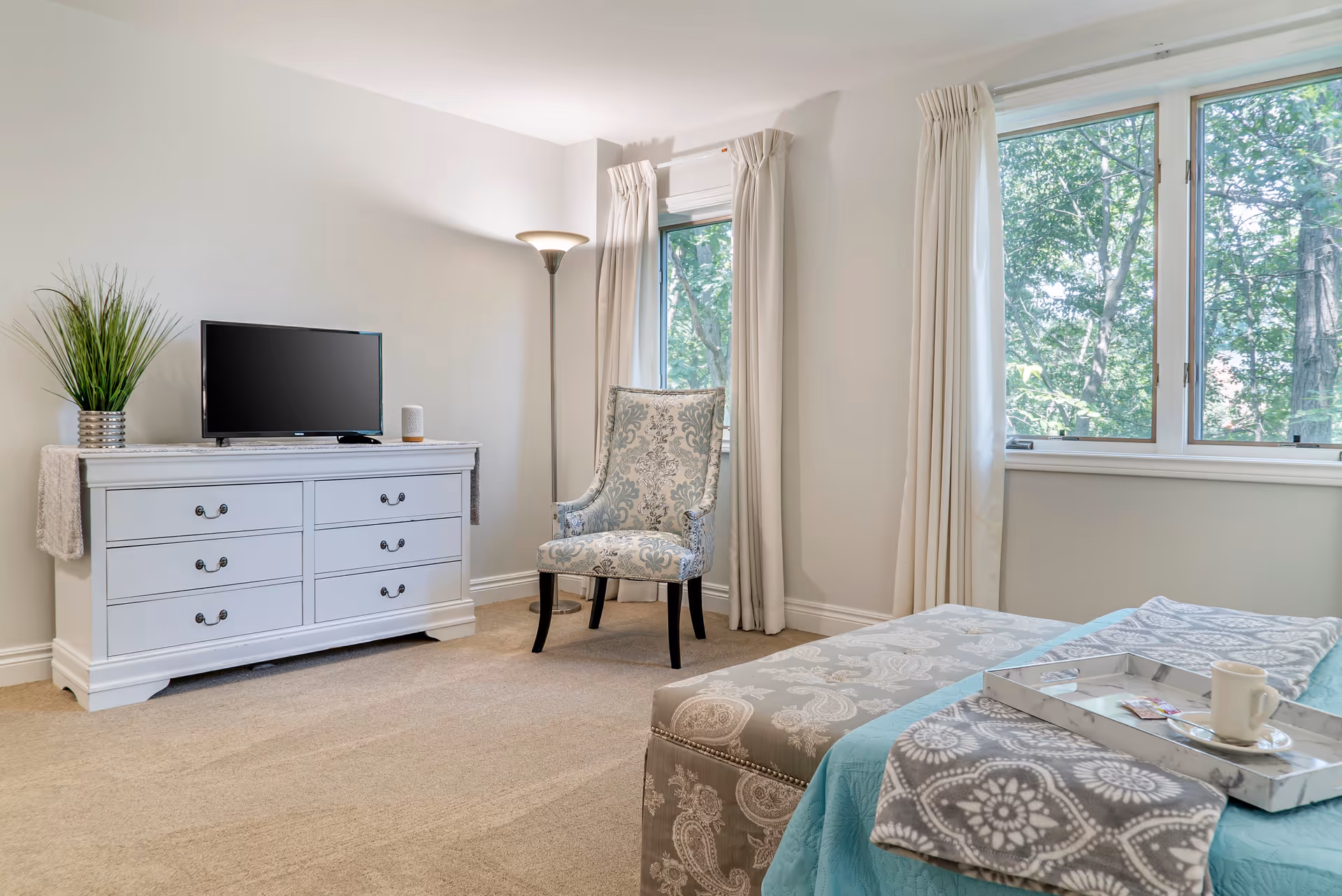 A bright bedroom with beige carpet, a white dresser holding a flat-screen TV and a small speaker, a patterned armchair near two windows with white curtains, and a bed with a blue and gray patterned blanket and a tray holding a cup and some packets.