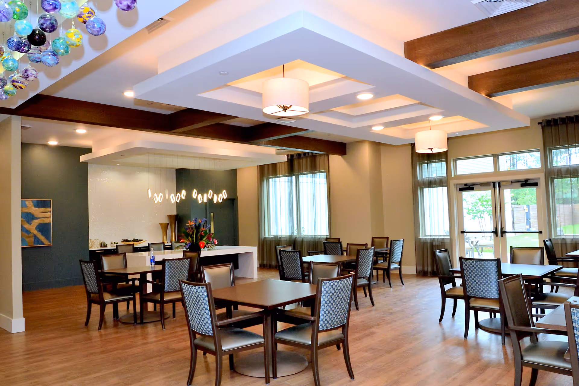 A spacious and well-lit dining area with multiple square tables and cushioned chairs arranged neatly. The room features large windows with sheer curtains allowing natural light to enter. Modern ceiling lights and decorative hanging glass orbs add to the ambiance. There is a counter area in the back with pendant lights and a flower arrangement.
