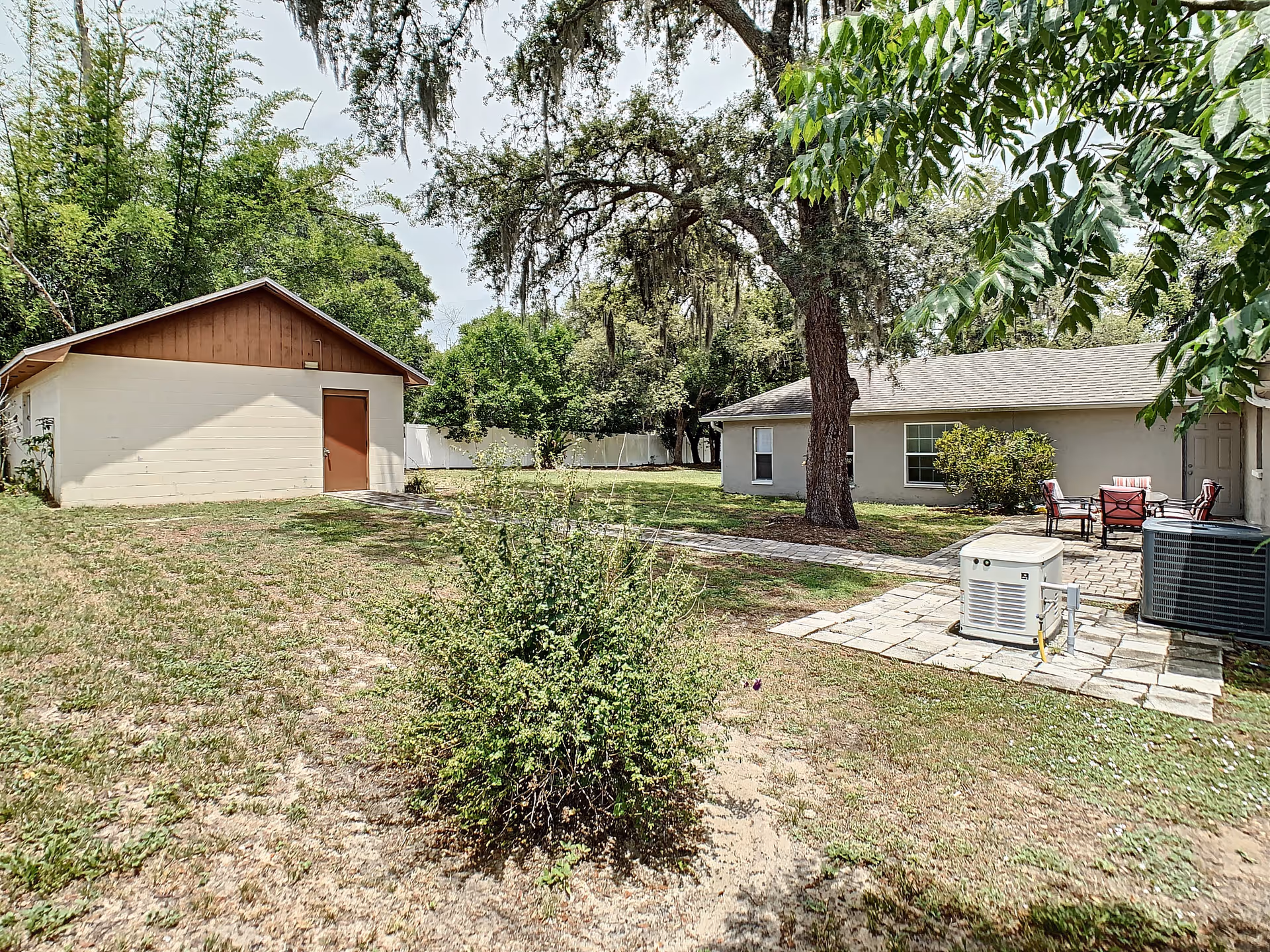 Outdoor view of a senior living facility showing a grassy yard with a small bush in the foreground, a large tree with hanging moss, a paved walkway, a small building with a brown door and roof on the left, and a beige building with windows and a patio area with chairs and a table on the right.
