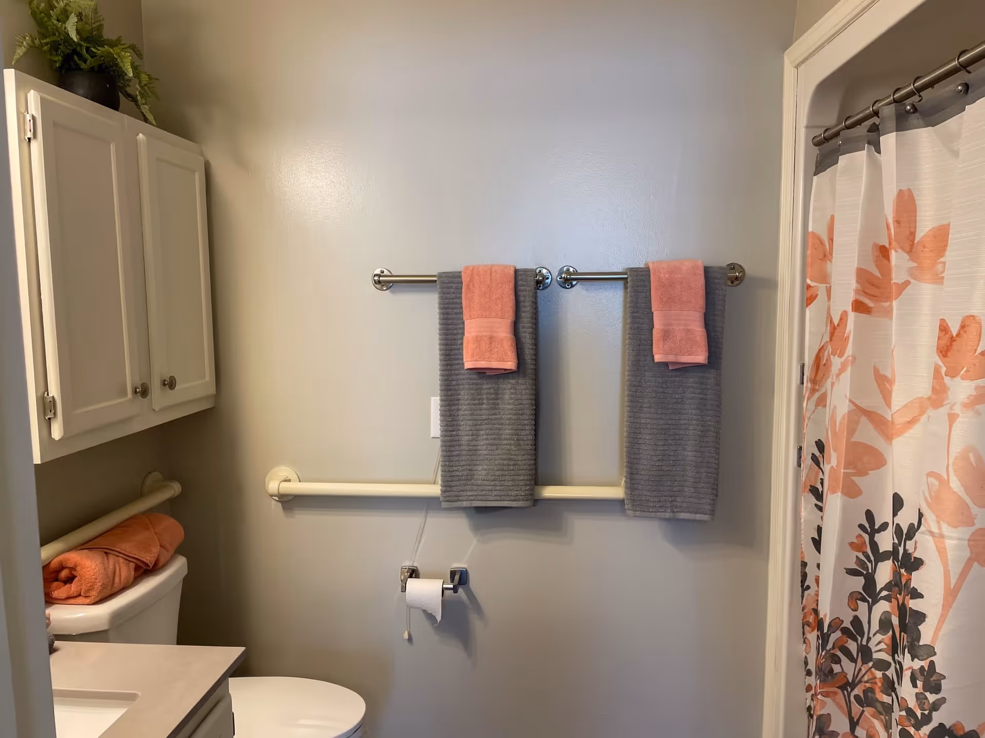 A bathroom with a white toilet, a white cabinet above it, and a countertop with a sink partially visible. Two gray towels with smaller pink towels on top hang on two towel bars mounted on a light gray wall. A white grab bar is installed horizontally on the wall behind the toilet and towels. To the right, a shower curtain with a floral pattern in shades of pink, orange, and black is visible.