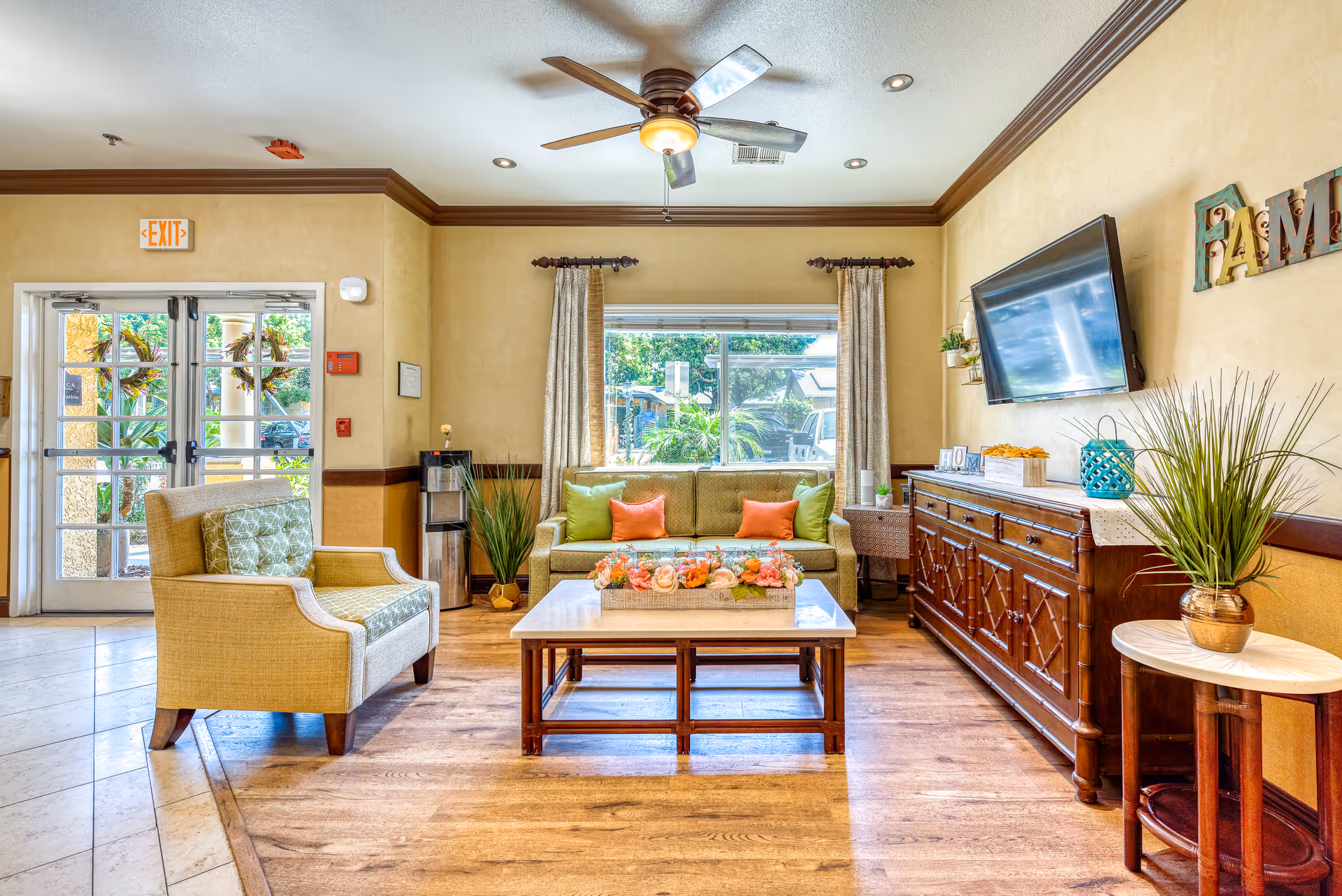 Sunlit communal living room with a sofa, armchair, coffee table, wall-mounted TV, and decorative plants.