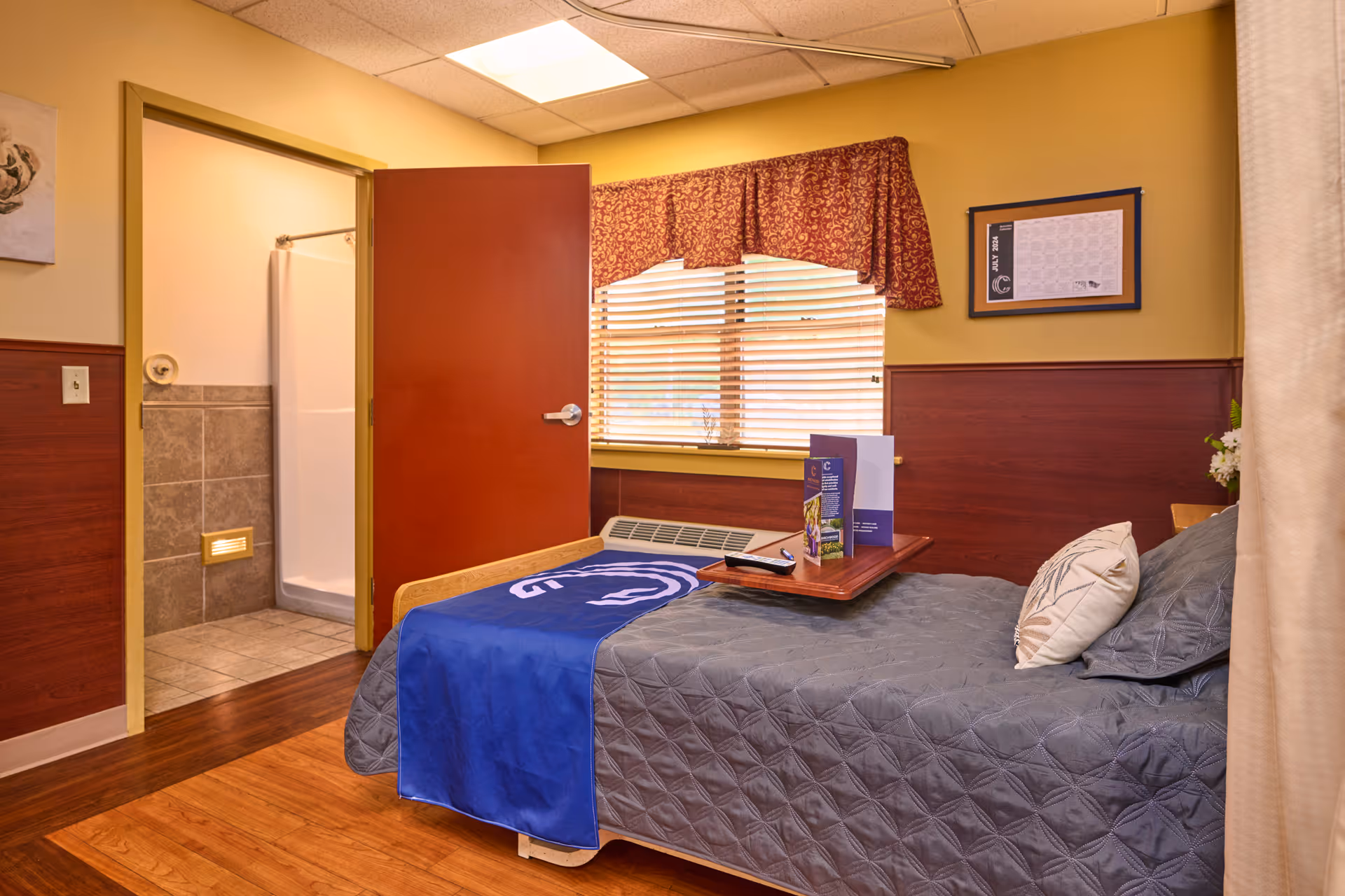 A cozy bedroom in a senior living facility with a single bed covered in a gray quilt and a blue blanket. There is a wooden tray on the bed holding a remote control and some brochures. The room has wooden flooring, a window with blinds and a red patterned valance, and a door leading to a bathroom with a shower stall. The walls are painted yellow with a wooden wainscot panel.