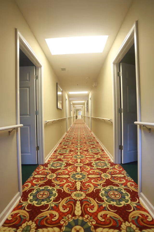 A long interior hallway in an assisted living facility with a patterned red carpet, handrails, and open doorways.