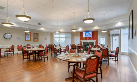 Spacious communal dining room with round tables, red-upholstered chairs, pendant lights, a fireplace with a mounted TV, and large windows.