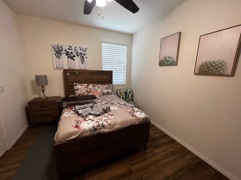 Small bedroom with a floral-patterned bed, wooden headboard and nightstand, wall art, a window with blinds, and a ceiling fan.