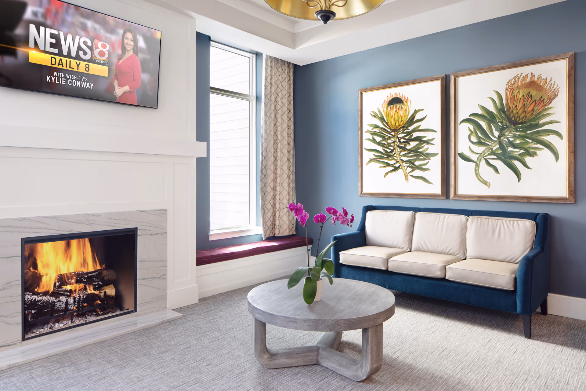 A cozy living room with a blue sofa featuring cream cushions, a round wooden coffee table with a purple orchid plant, a lit fireplace with a white and gray marble surround, a window with patterned curtains and a cushioned window seat, and two large botanical paintings on a blue wall. A flat-screen TV above the fireplace displays a news program.