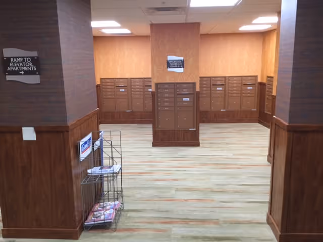 Interior view of a mailroom area with multiple brown mailboxes mounted on the walls and a central mailbox unit. The room has wood-paneled lower walls, orange upper walls, and a light-colored floor with subtle patterns. There is a metal rack with magazines and a sign indicating a ramp to elevator apartments.