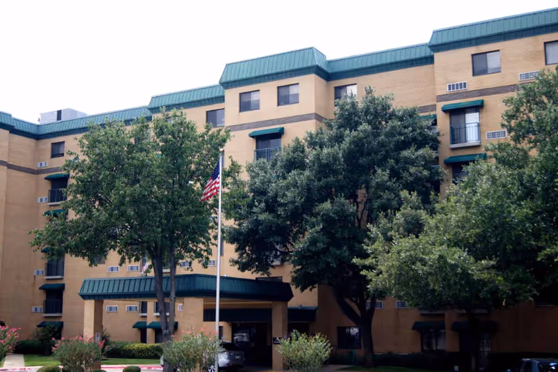 Exterior view of a multi-story brick building with green awnings and a green roof, surrounded by trees and bushes. An American flag is flying on a flagpole in front of the building.