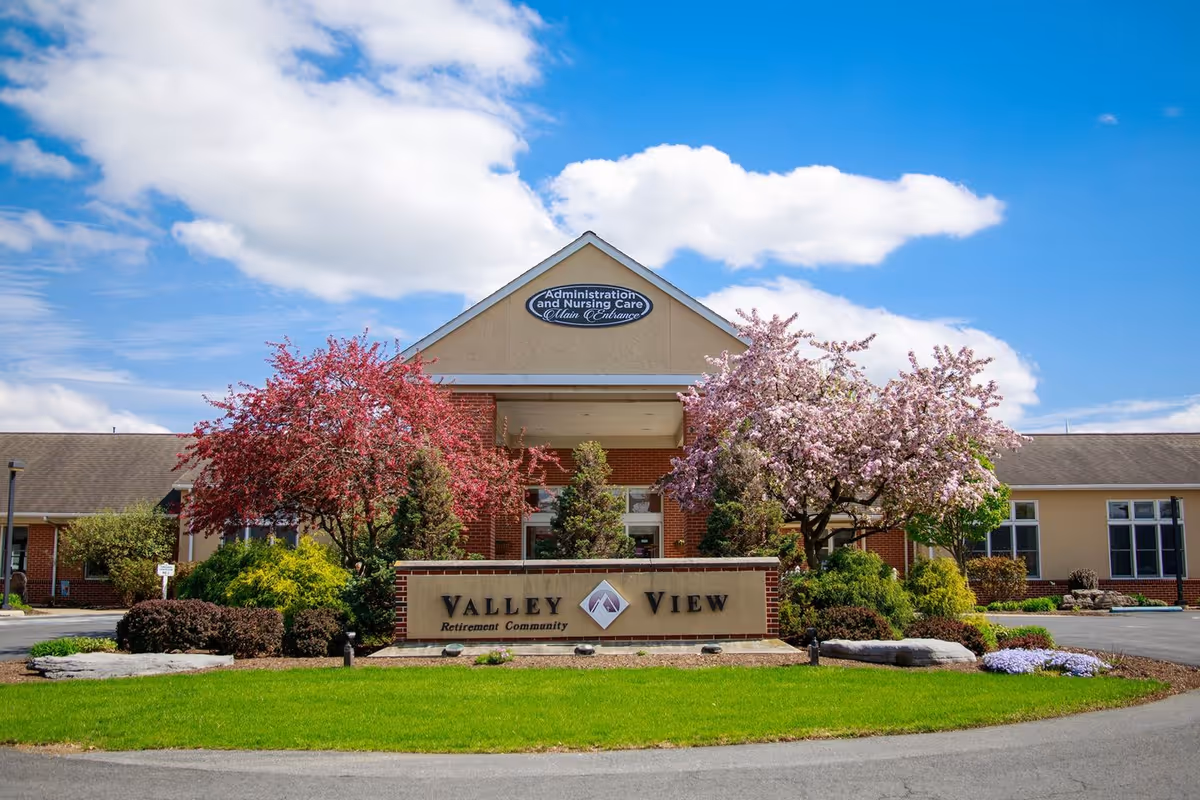 Front exterior view of Valley View Retirement Community building with a sign in front, surrounded by green grass, bushes, and blooming pink and red trees under a blue sky with some clouds.