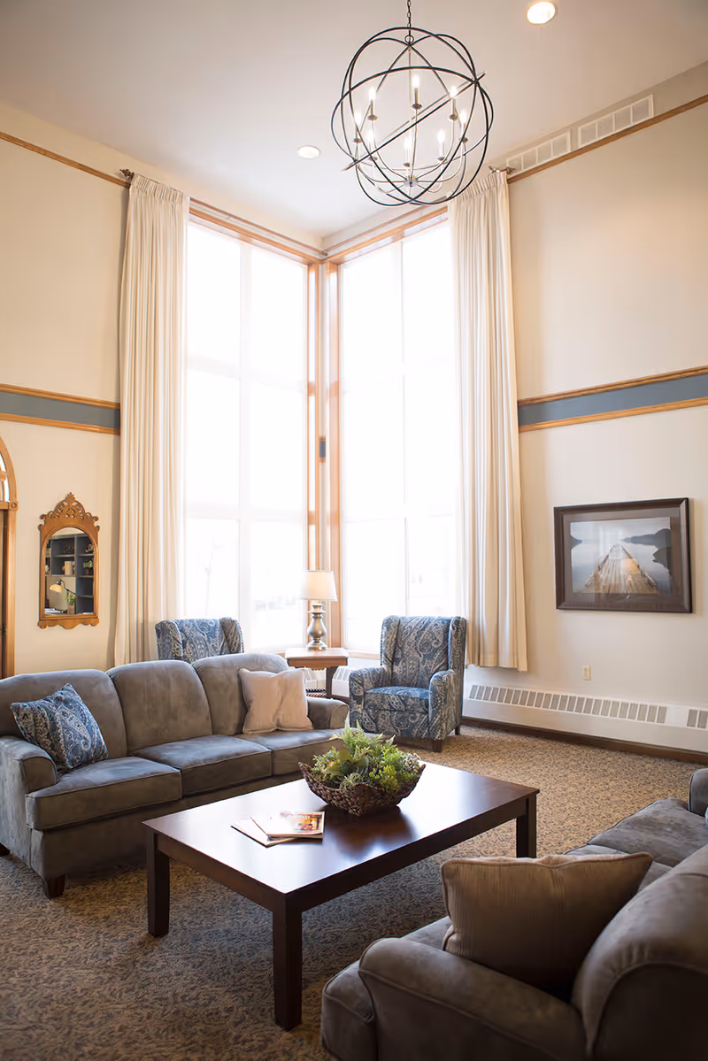 A bright and spacious living room with large floor-to-ceiling windows covered by cream-colored curtains. The room features a gray sofa and armchair with decorative pillows, two patterned blue armchairs, a wooden coffee table with a plant centerpiece and magazines, a side table with a lamp, a framed picture on the wall, and a decorative mirror. The ceiling has a modern spherical chandelier.