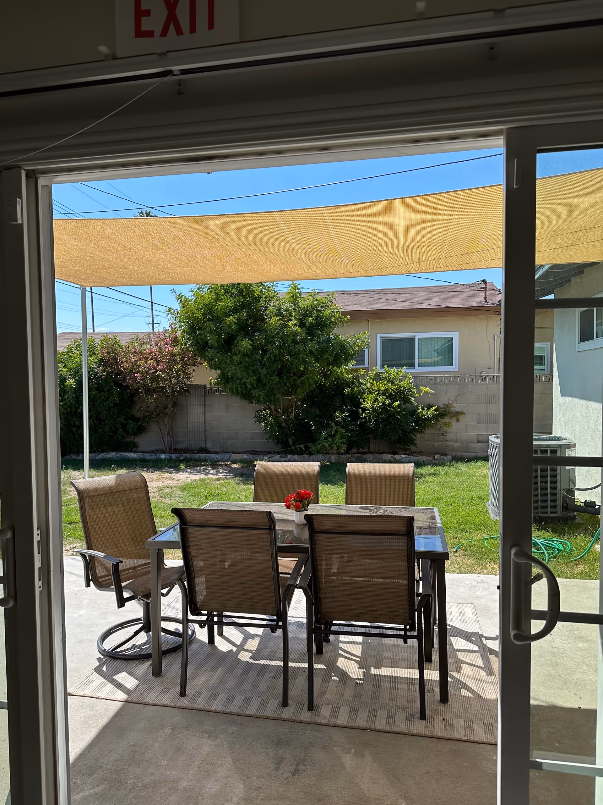 View through sliding glass doors of a backyard patio with a dining table and six chairs under a tan shade sail and a small lawn with shrubs.