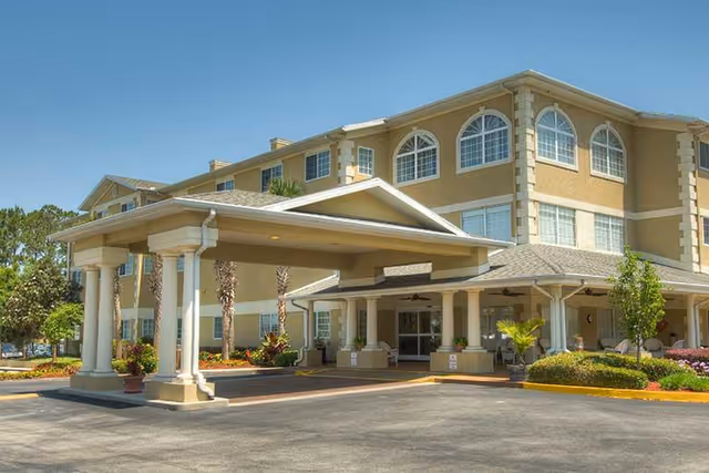 Exterior view of a multi-story senior living facility building with a covered entrance supported by columns, surrounded by landscaped greenery and a clear blue sky.