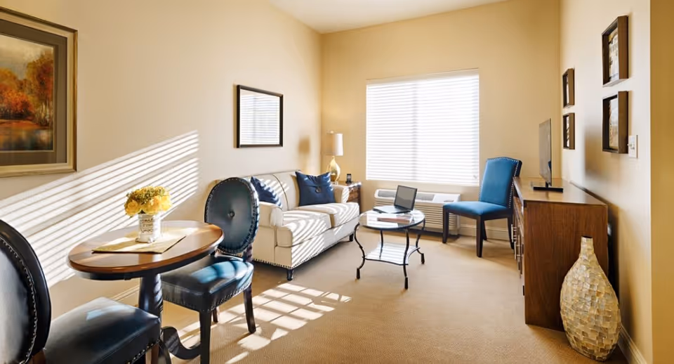 A bright living room with beige walls and carpet, featuring a cream-colored sofa with blue pillows, a small round glass coffee table, a wooden TV stand with a flat-screen TV, a blue upholstered chair, and a small round wooden table with two blue chairs. Sunlight streams through a window with blinds, casting shadows on the walls and floor. Decorative artwork and a large vase are also visible.