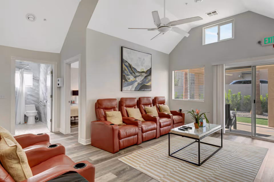 A bright living room with four red leather recliner chairs arranged in a row, each with a beige pillow. A modern coffee table with a plant and remote controls sits on a striped rug. The room has high vaulted ceilings with a ceiling fan, a large window, and sliding glass doors leading to an outdoor patio. To the left, there is a doorway leading to a bathroom with a toilet and shower visible, and another doorway leading to a bedroom.