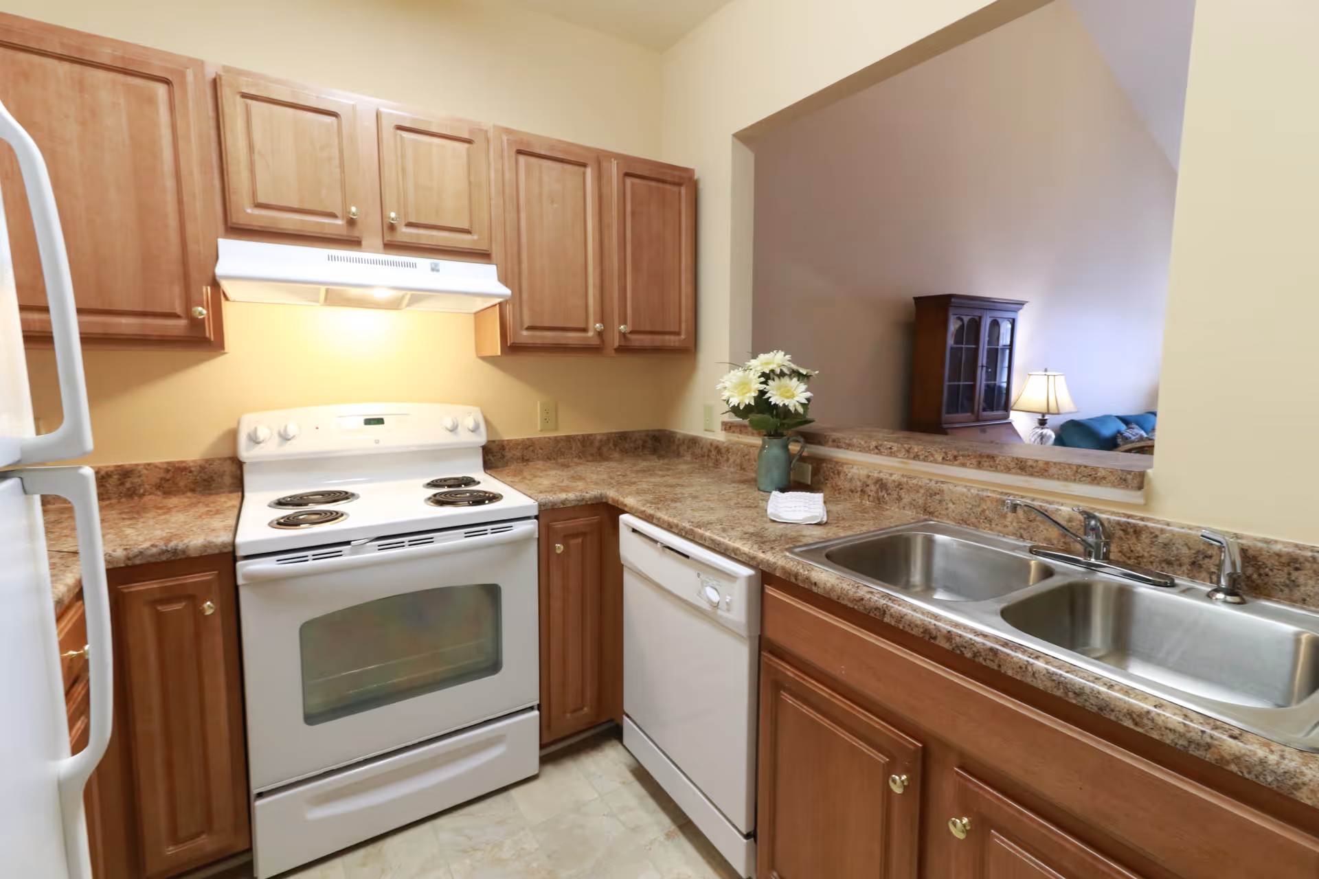 A small kitchen with a white stove, dishwasher and double sink set in brown laminate countertops and wood cabinets, open to a living area.