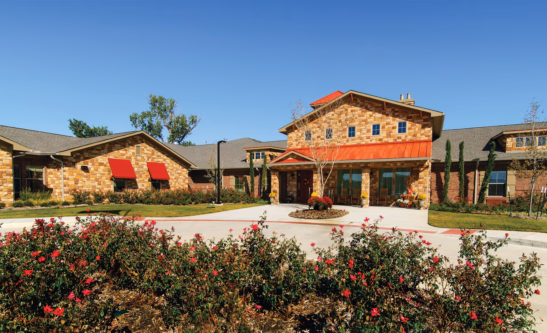 Exterior view of a single-story assisted living and memory care facility with stone and brick facade, red awnings, a red metal roof over the entrance, and landscaped grounds with bushes and flowers in front.