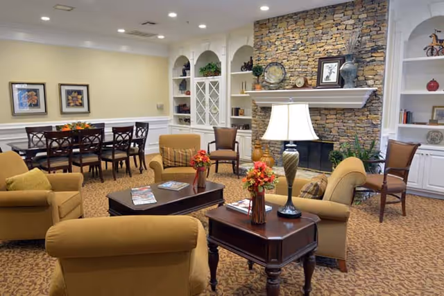 A cozy senior living common area with beige armchairs and a wooden coffee table adorned with flower vases and magazines. The room features a stone fireplace with decorative items on the mantel, built-in white shelves with plants and ornaments, and a dining table with chairs in the background. The carpet is patterned and the walls are painted light yellow and white.