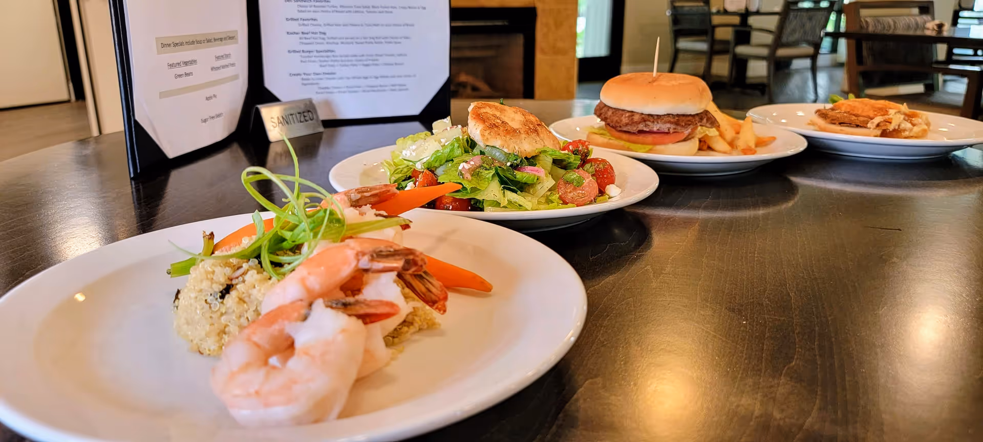 A close-up view of a dining table with four plates of food including shrimp with quinoa and vegetables, a fresh salad with a biscuit, a hamburger with fries, and a dessert. In the background, there is a menu and a small sign that says 'SANITIZED'.