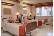 A bright and clean bedroom in a senior living facility with two single beds, each with white bedding and red throws. The room features bedside tables with lamps and decorative plants, a large window with blinds and a valance, and a neutral color scheme with warm accents.