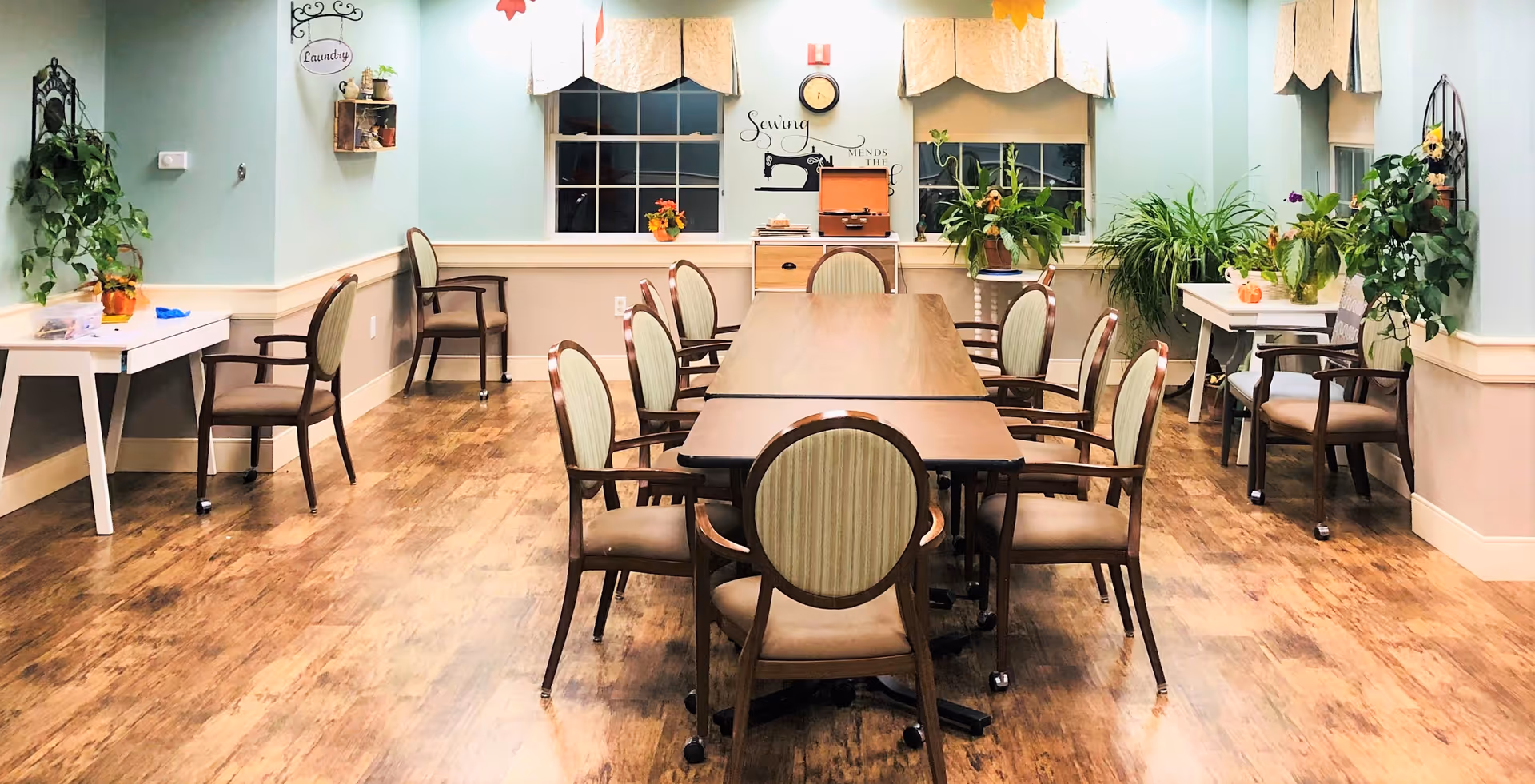 A well-lit room with a long table surrounded by chairs, decorated with various green plants on side tables and windowsills. The walls are painted light blue with beige wainscoting, and there are two windows with beige valances. A wall decoration features a sewing machine and the phrase 'Sewing mends the soul.'