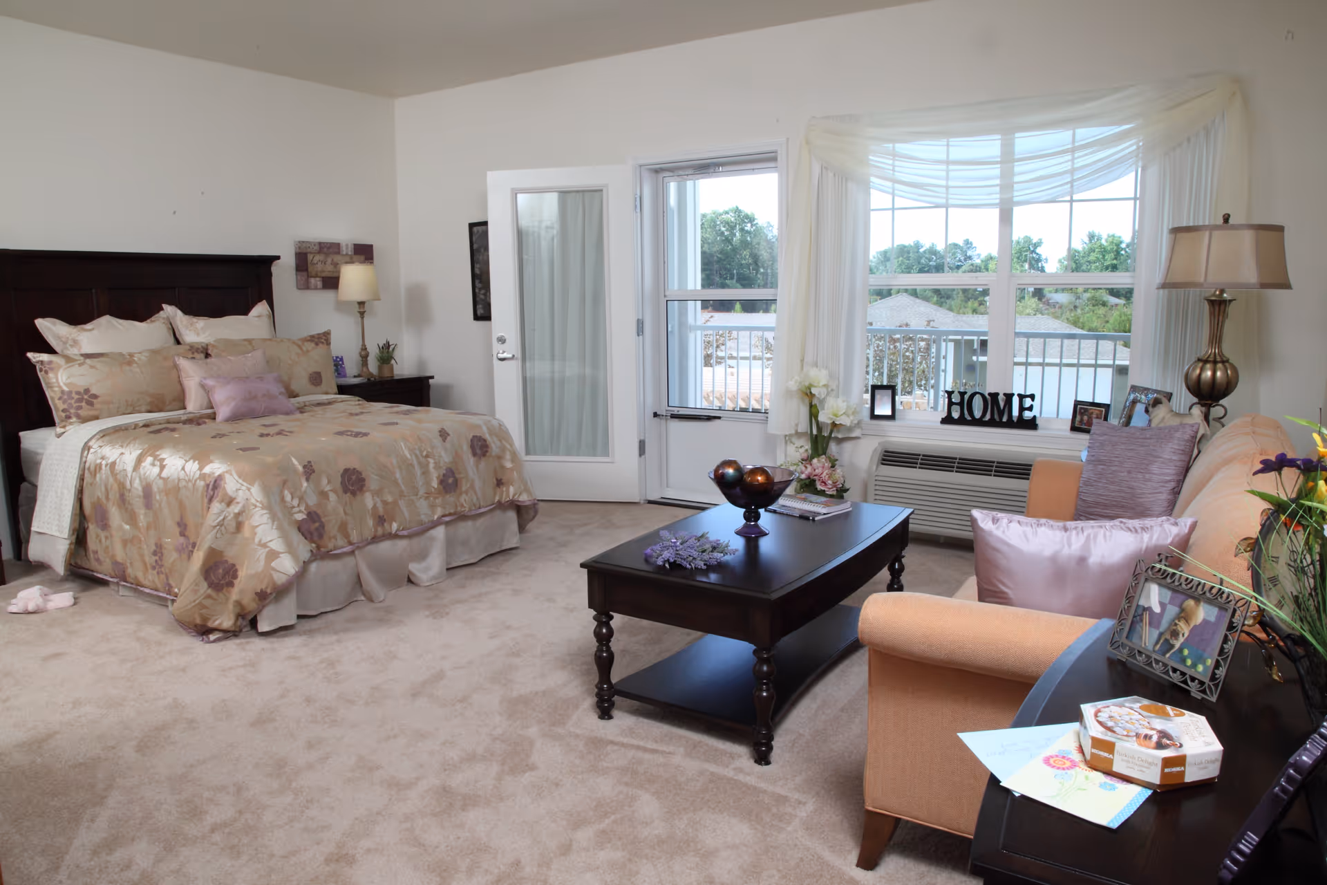 A cozy bedroom with a large bed dressed in floral bedding, a wooden headboard, and multiple pillows. Next to the bed is a nightstand with a lamp and small decorative items. The room has beige carpet and a glass door leading to a balcony. In front of a large window with sheer curtains, there is a peach-colored sofa with purple pillows, a dark wooden coffee table with decorative items, and a side table with framed photos and a box of snacks.