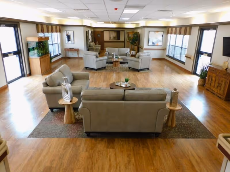 Spacious senior living common area with multiple beige sofas and armchairs arranged on rugs in a large room with wood floors and windows.