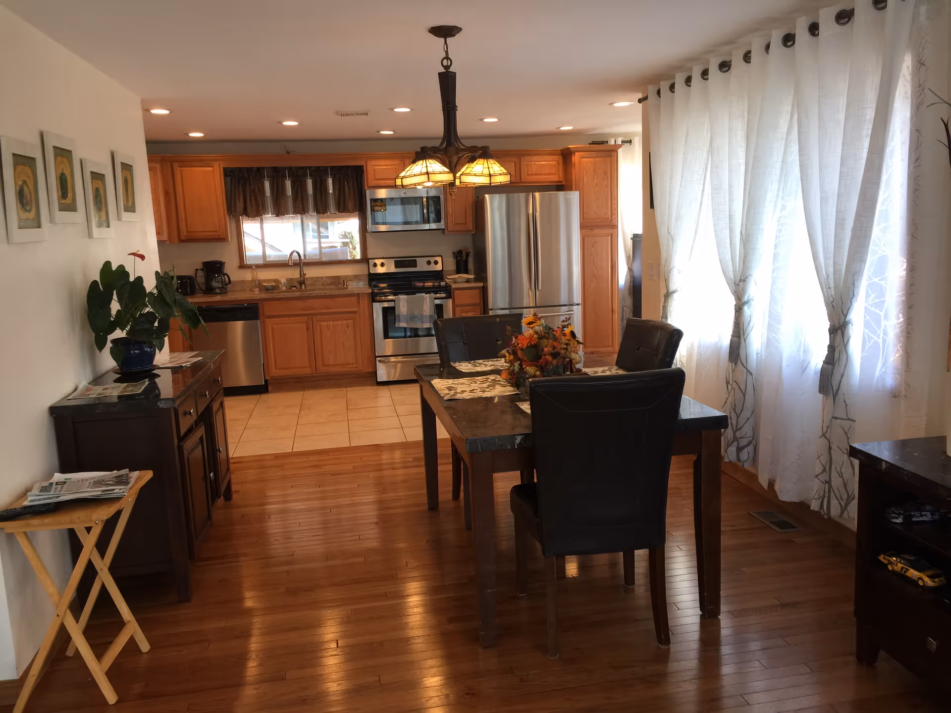 A cozy kitchen and dining area with wooden floors and cabinetry. The kitchen features stainless steel appliances including a refrigerator, oven, microwave, and dishwasher. A wooden dining table with four black cushioned chairs is centered in the room, decorated with a floral centerpiece. Large windows with sheer white curtains allow natural light to fill the space. The walls are adorned with framed artwork and there is a small side table with a plant and magazines.