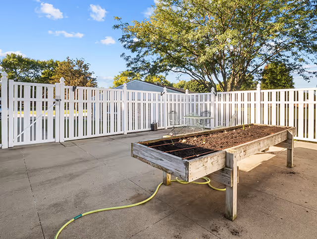 A sunlit outdoor courtyard with a raised wooden planter bed, white vinyl fence, patio furniture, and a yellow garden hose.