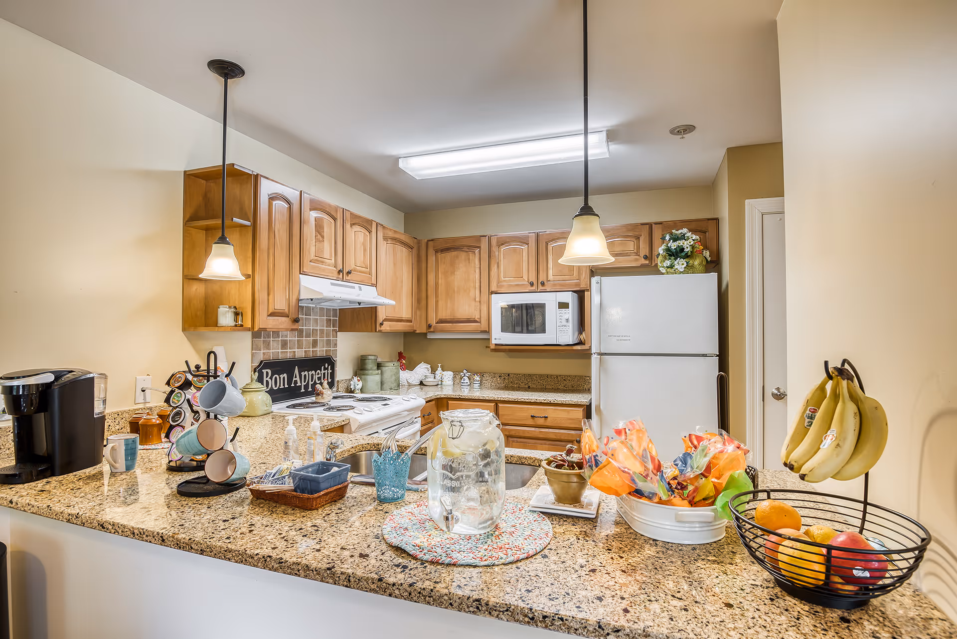 A well-lit kitchen with wooden cabinets, granite countertops, and various kitchen items including a coffee maker, mugs on a rotating rack, a glass water dispenser, fruit basket with bananas and other fruits, and a white refrigerator. Two pendant lights hang over the counter, and a 'Bon Appetit' sign is visible near the stove.