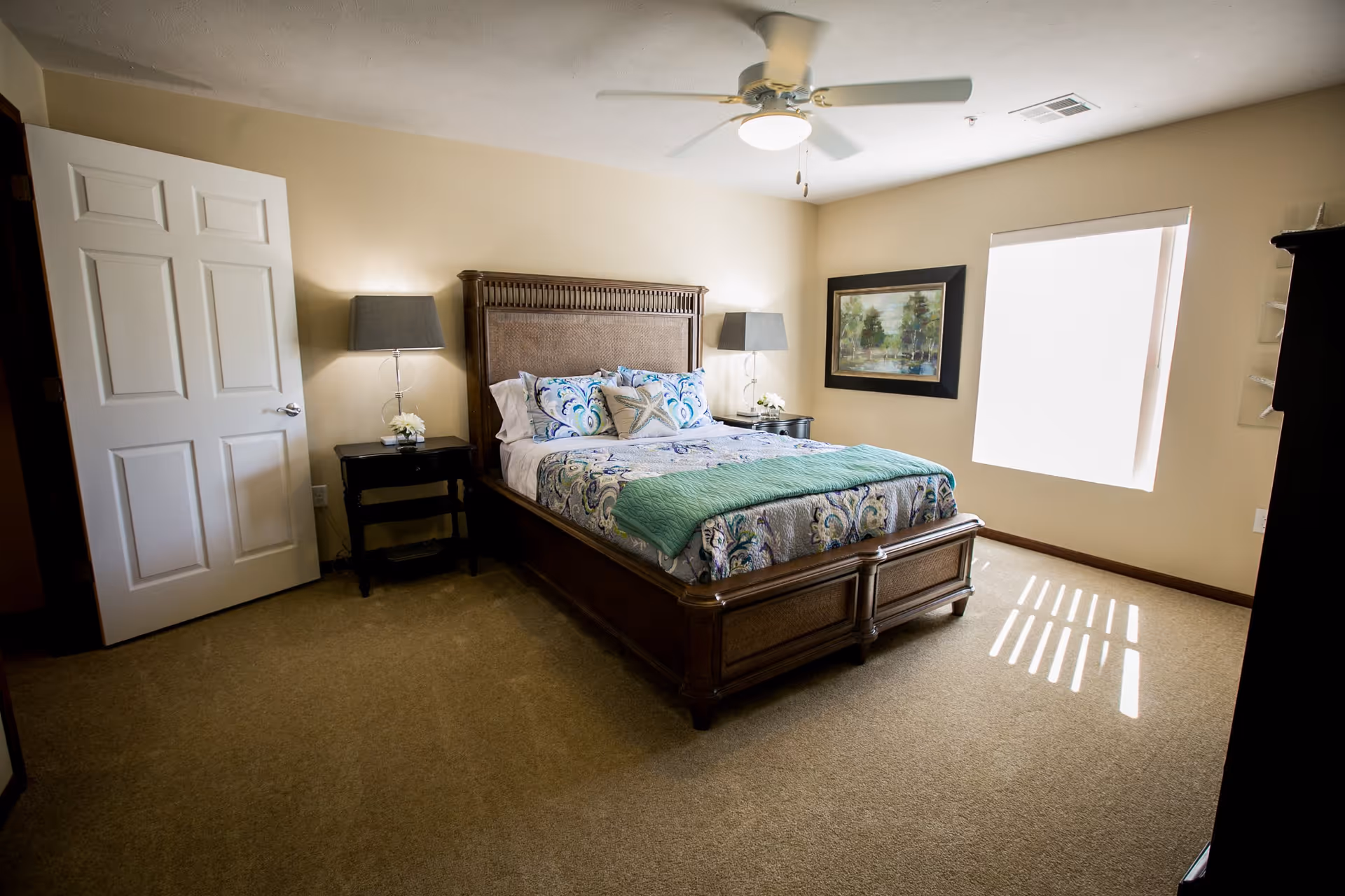A well-lit bedroom with a large wooden bed featuring a patterned bedspread and decorative pillows. There are two bedside tables with lamps and small flower arrangements. A ceiling fan is mounted above, and a framed painting hangs on the wall next to a window with blinds partially open, allowing sunlight to cast shadows on the carpeted floor.