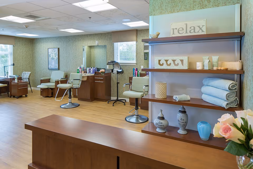 Bright salon-style interior with styling chairs, shelving holding towels and decor, and a reception desk.