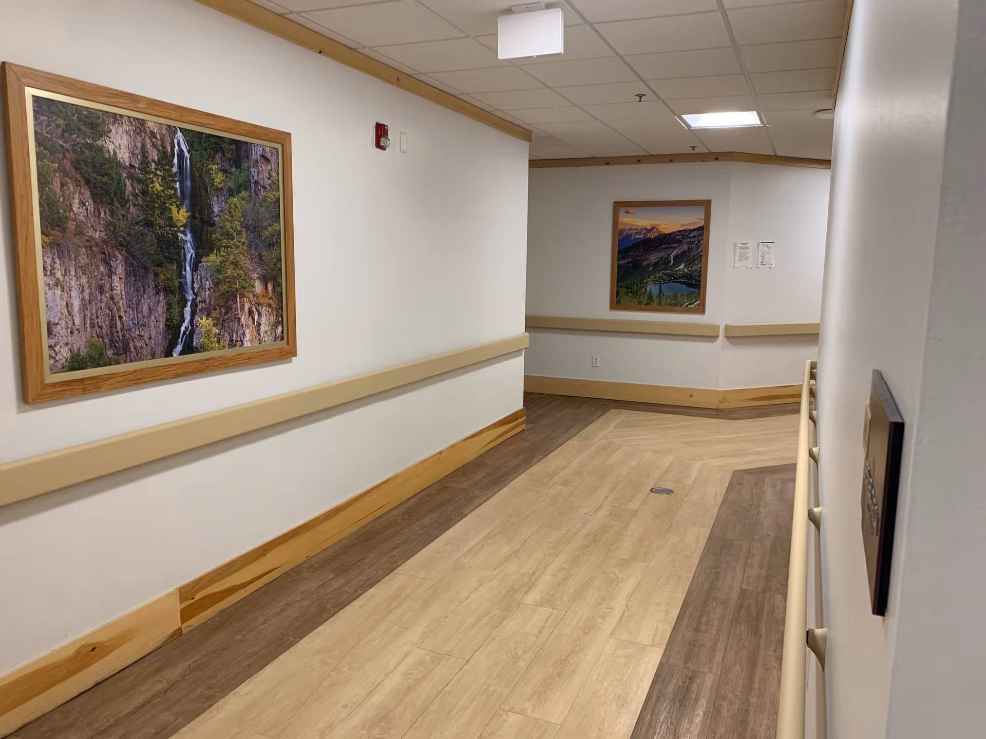 A clean, well-lit hallway in a nursing and rehabilitation facility with light-colored walls and wood-tone flooring. The hallway features handrails on both sides and framed landscape photographs hanging on the walls. The ceiling has recessed lighting panels.