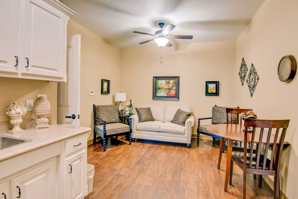 A cozy interior sitting area with a white sofa, two chairs, a small dining table, and a kitchenette corner under a ceiling fan.