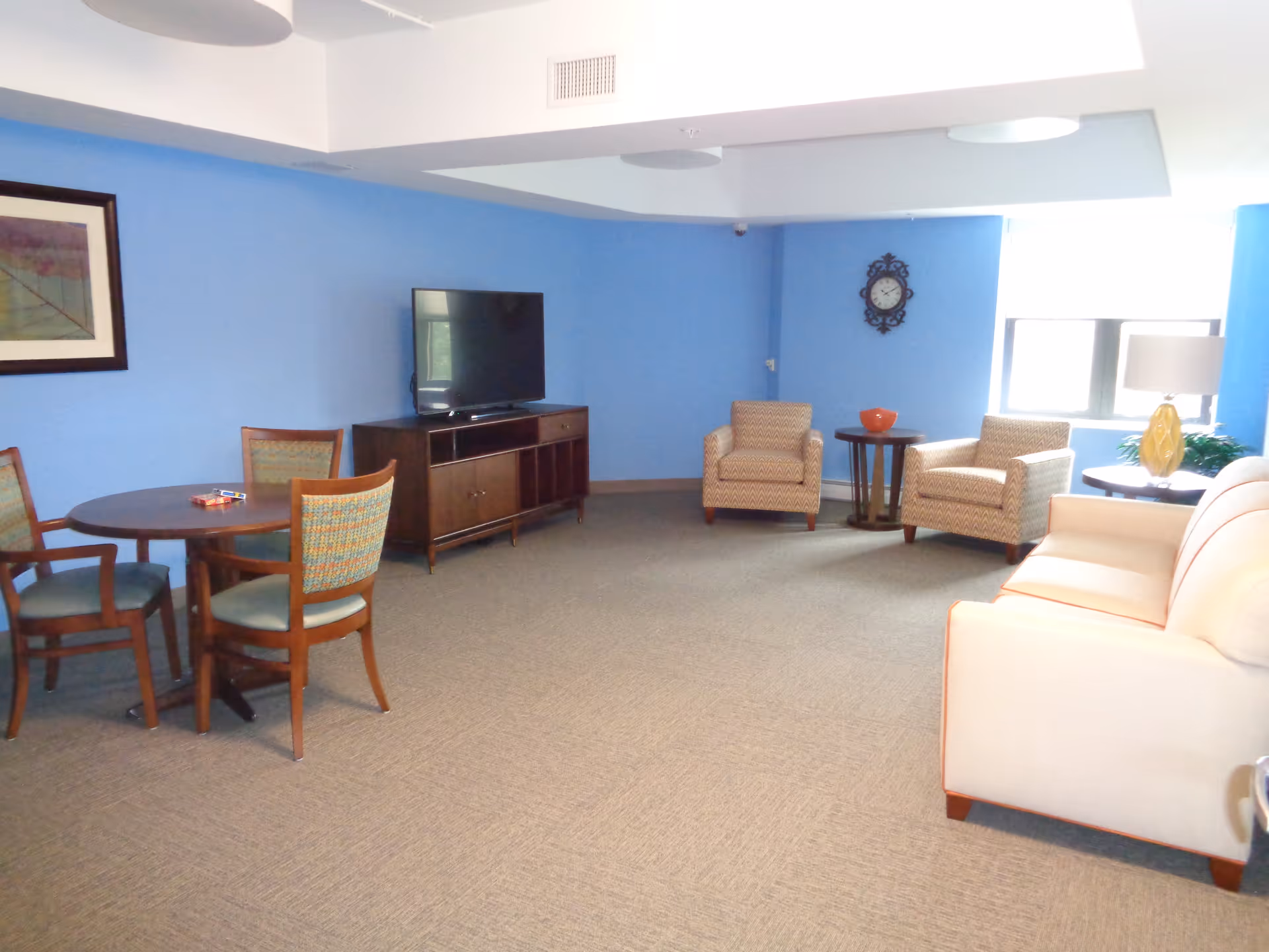 A spacious living room with light blue walls and beige carpet. The room contains a beige sofa, two patterned armchairs, a round wooden table with four chairs, a wooden TV stand with a flat-screen TV, a side table with a lamp, a wall clock, and a framed picture on the wall.