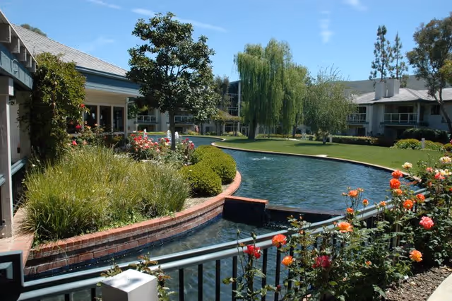 A landscaped outdoor area at Chateau Lake San Marcos featuring a winding water canal bordered by a brick edge, lush green grass, various trees including a willow, and colorful blooming flowers. Residential buildings with balconies surround the garden under a clear blue sky.