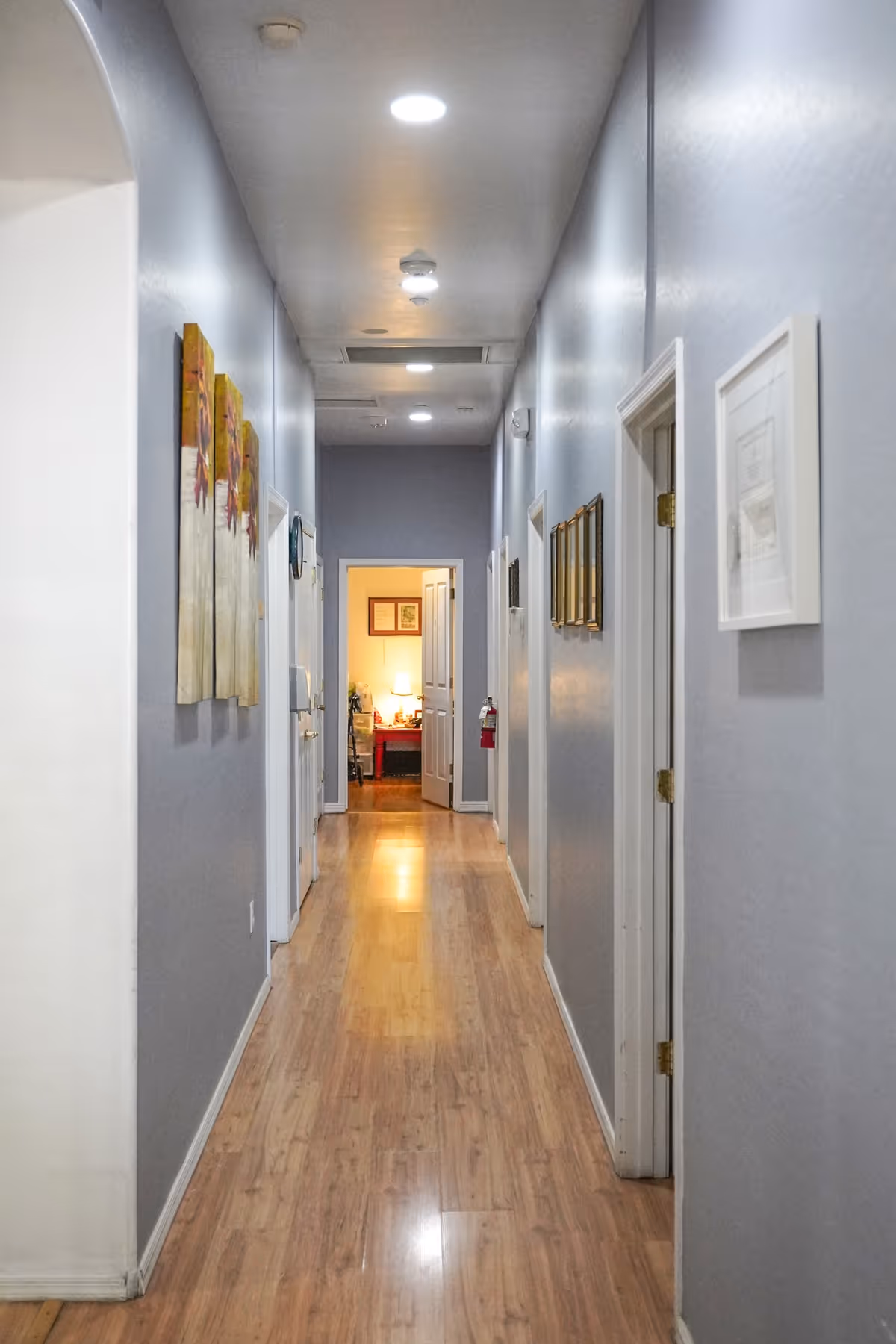A long, narrow hallway with light wooden flooring and gray walls. Several doors line both sides of the hallway, and framed artwork is hung on the walls. The hallway is well-lit with recessed ceiling lights, and at the far end, an open door reveals a warmly lit room with a table lamp and framed pictures on the wall.