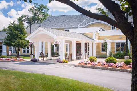 Front entrance of The Arbors of Bedford with white portico, colorful flower beds, and landscaping.