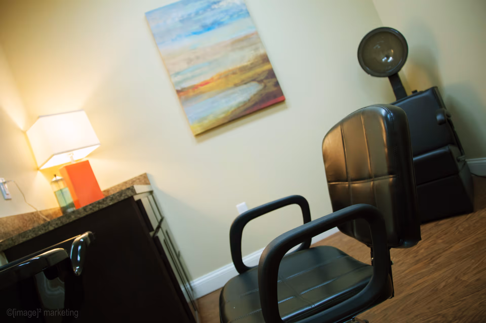 Interior view of a room with a black salon chair in the foreground, a lamp on a countertop to the left, a colorful abstract painting on the wall, and a hair dryer chair in the background.