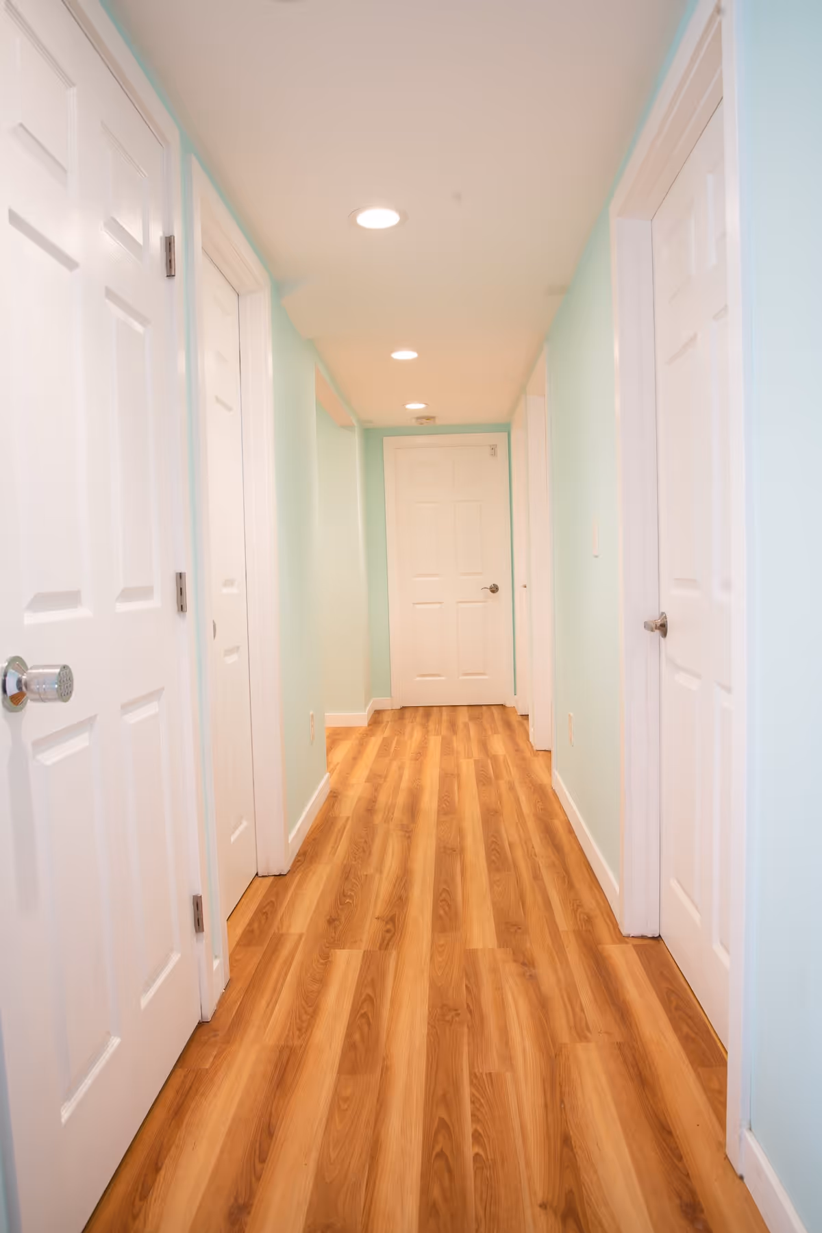 A well-lit hallway with light blue walls and wooden flooring. The hallway has multiple white doors on both sides and recessed ceiling lights.