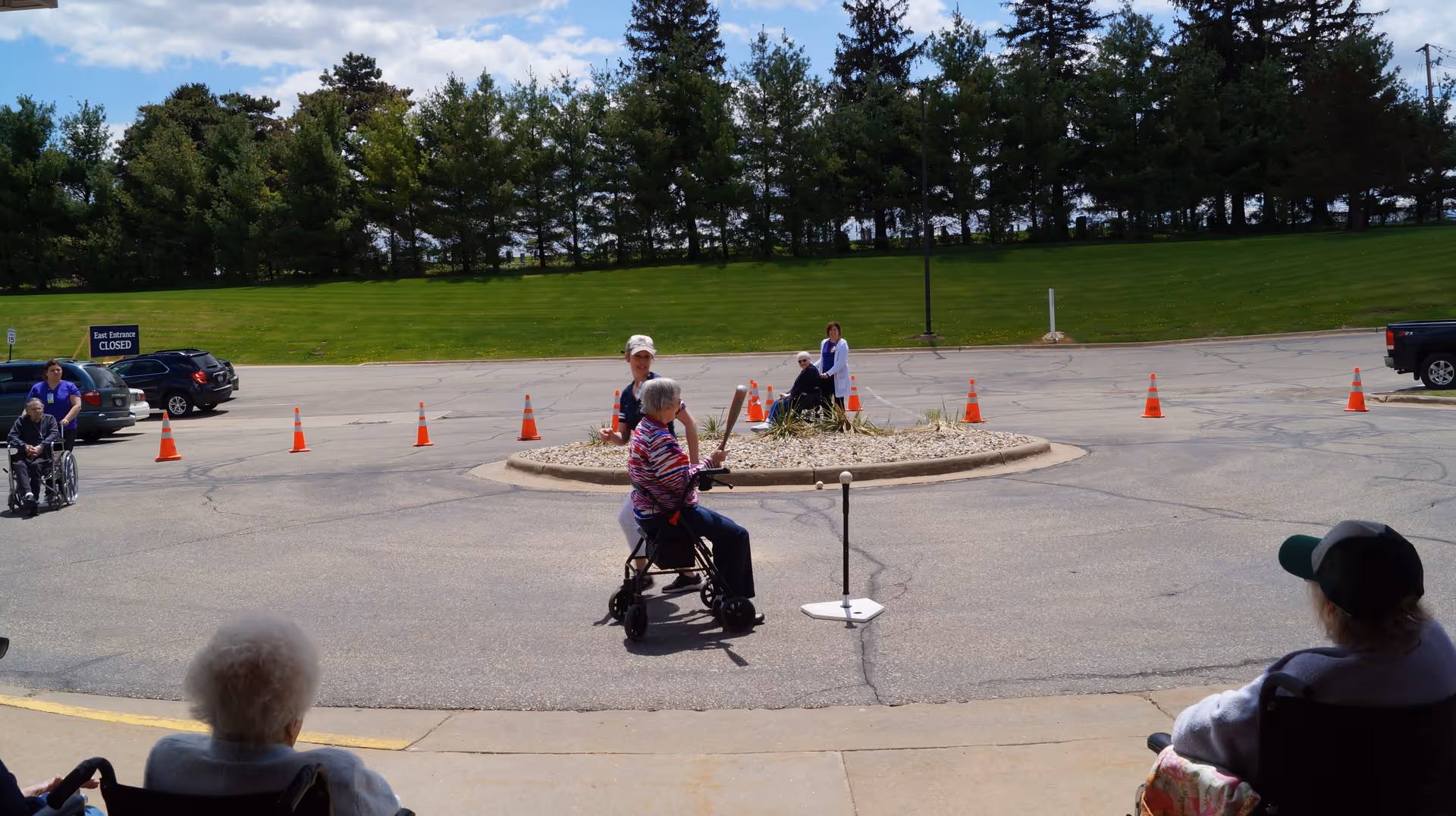 Several elderly individuals, some in wheelchairs, are outside in a parking lot area near a circular island with plants. Orange traffic cones are set up around the island. The background shows a grassy hill with trees and a clear sky.
