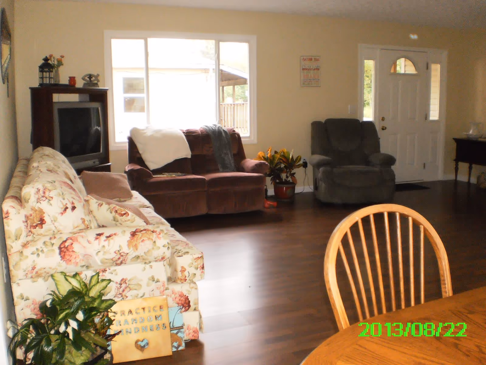 A living room with a floral patterned sofa, a brown loveseat with blankets draped over it, a gray recliner, a wooden TV stand with an older model television, a wooden dining table with chairs, and a white front door with windows on either side. There are plants and decorative items placed around the room, and a large window letting in natural light.