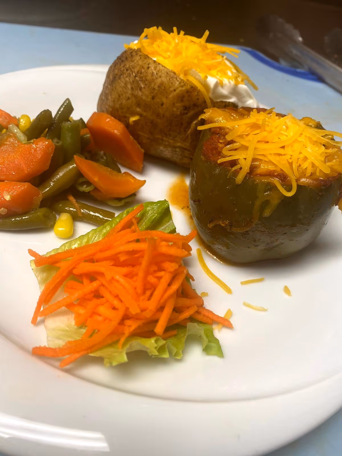 A plate of food featuring a baked potato topped with shredded cheddar cheese and sour cream, a stuffed green bell pepper topped with shredded cheddar cheese, cooked mixed vegetables including green beans, carrots, and corn, and a small salad with shredded carrots on a lettuce leaf.