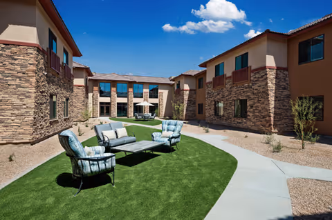 Outdoor courtyard area of a senior living facility with artificial grass, outdoor seating including cushioned chairs and a sofa, surrounded by two-story buildings with stone and stucco exteriors under a blue sky with scattered clouds.