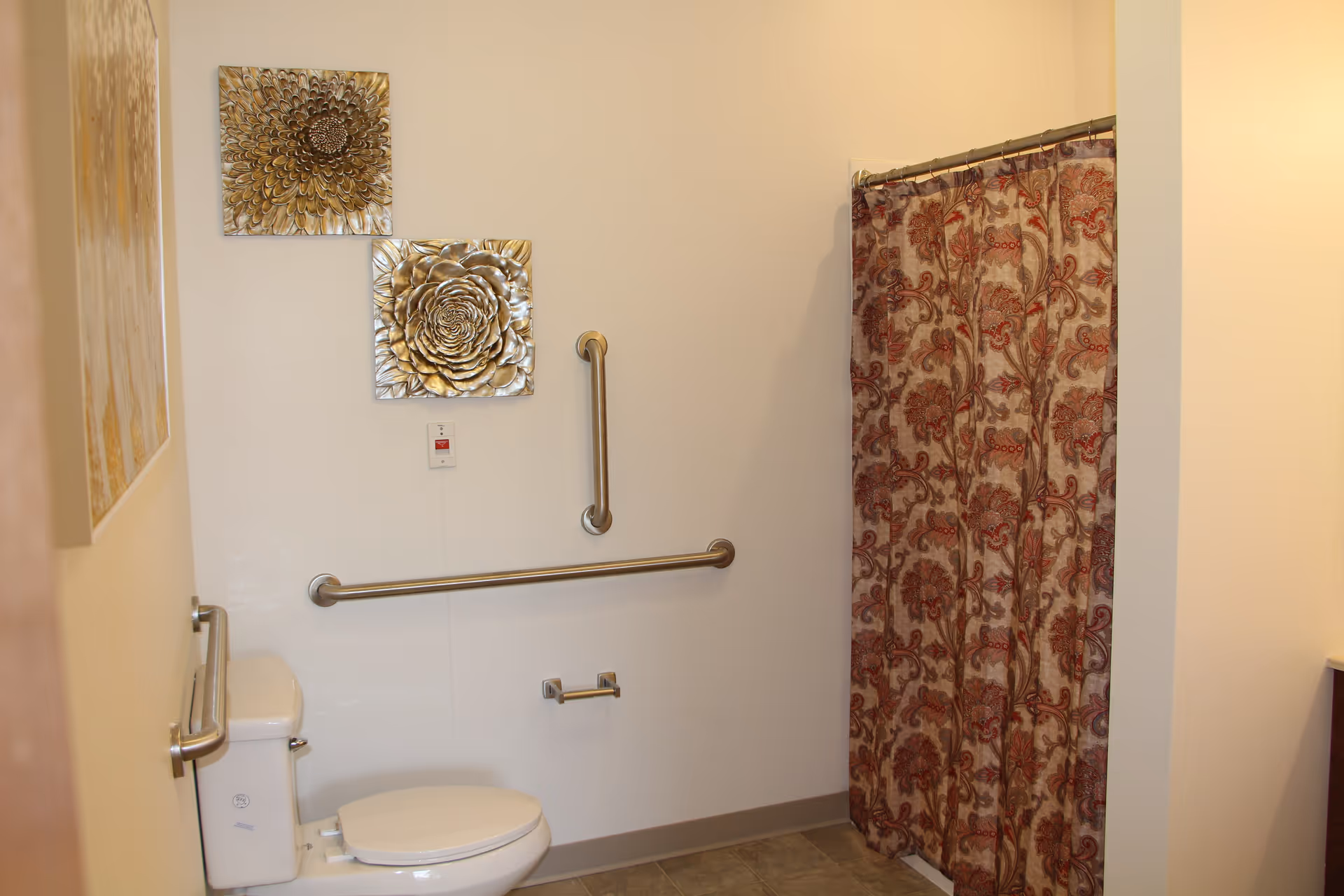 A senior living facility bathroom with a white toilet, two metal grab bars on the walls, and a shower area with a floral patterned curtain. The walls are decorated with two metallic flower wall art pieces.