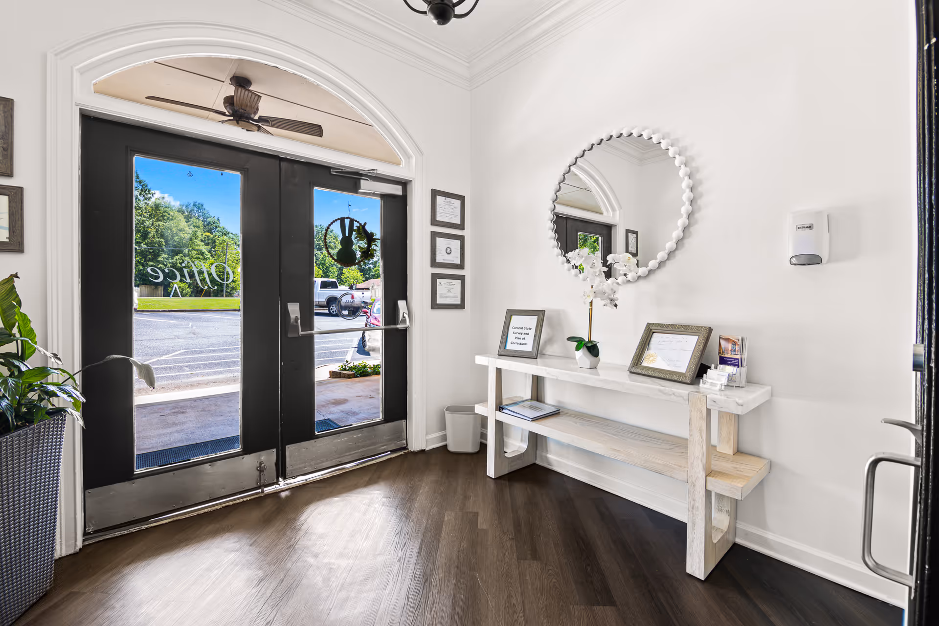 Entrance area of Avery Village Assisted Living and Memory Care featuring double glass doors with black frames and an arched window above. Inside, there is a white wooden bench with a white marble top holding framed certificates, a white orchid plant, and brochures. A round mirror with a beaded frame hangs on the wall above the bench. The floor is dark wood, and a hand sanitizer dispenser is mounted on the wall.