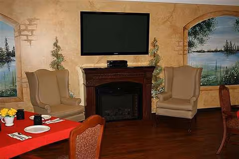 Living room with fireplace, two beige armchairs, a TV above mantel, and a red tablecloth.