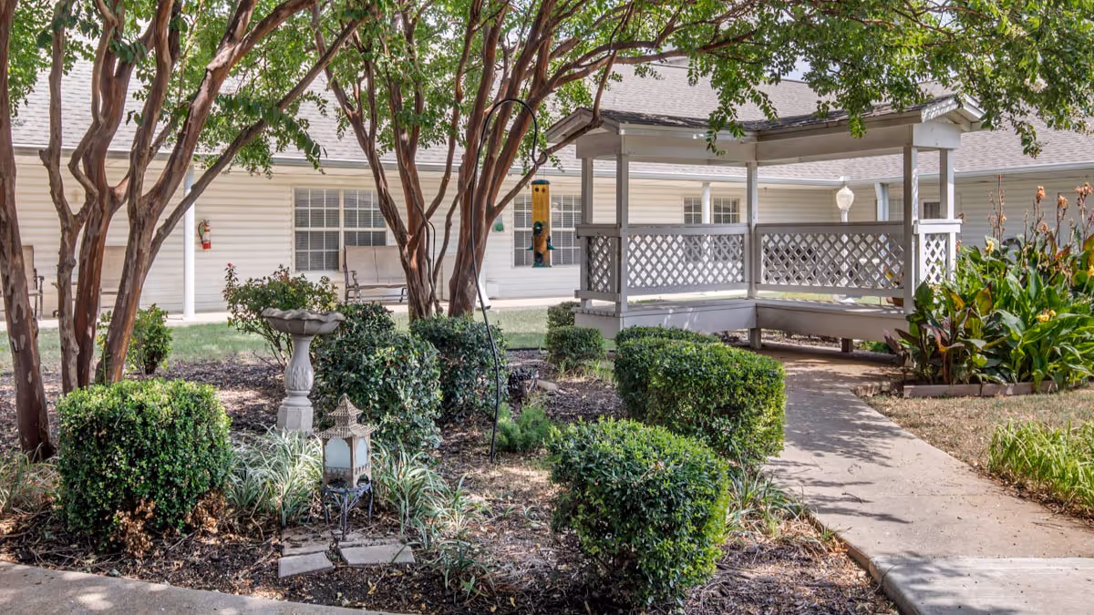 Outdoor garden area with trimmed bushes, trees, a birdbath, and a white gazebo with lattice railings. A concrete pathway leads to the gazebo, and a building with white siding and windows is visible in the background.