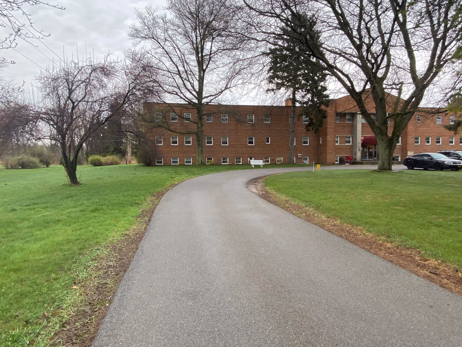 Curving driveway leading to the front of a three-story brick building surrounded by grass and trees.