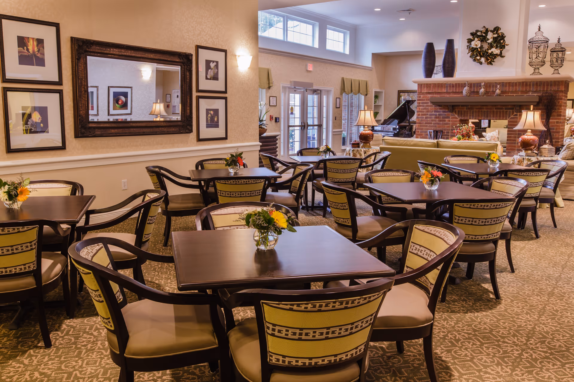 A cozy senior living dining area with multiple dark wood tables and cushioned chairs arranged neatly. Each table has a small vase with colorful flowers. The room features warm lighting, framed artwork on the walls, a large mirror, and a brick fireplace with decorative items on the mantel. Large windows and glass doors allow natural light to fill the space.